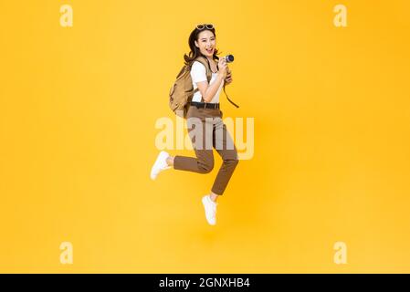 Giovane donna asiatica graziosa turista zaino in spalla sorridente e saltando con la macchina fotografica in mano isolato su sfondo giallo studio Foto Stock