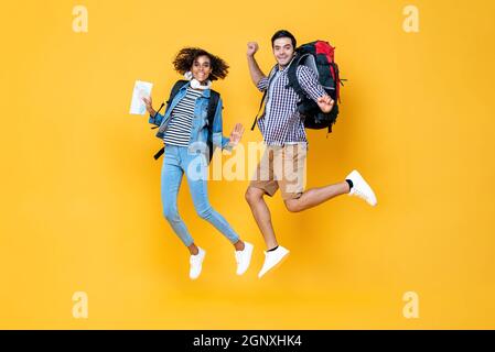 Felice giovane coppia turistica interrazziale con zaini pronti per viaggiare saltando in giallo sfondo di colore Foto Stock