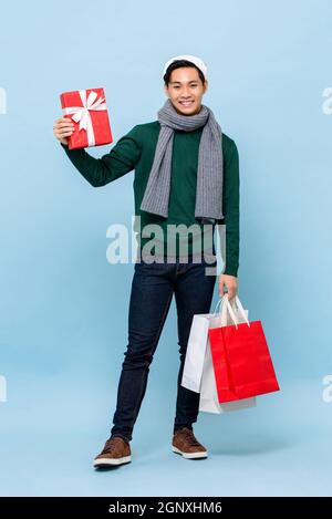Uomo asiatico felice che indossa Natale Santa cappello che tiene scatola regalo e borse shopping in blu chiaro studio isolato sfondo Foto Stock