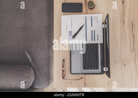 Piano di lavoro con scrivania da ufficio. Laptop, fattura e occhiali su tavolo di legno, divano. Foto Stock