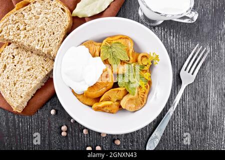 Zafferano di funghi salati con panna acida, foglia di curry e sprig di aneto in piatto, forchetta, tovagliolo e pane su sfondo di legno dall'alto Foto Stock