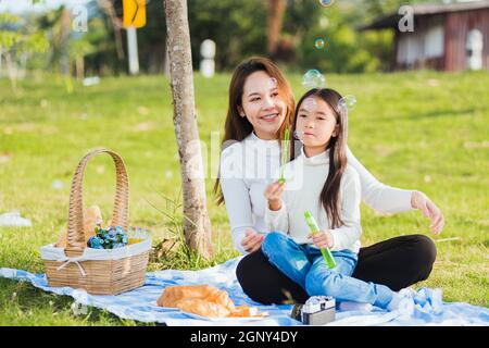 Buona madre asiatica e bambina bambina che si divertono e godendo all'aperto insieme seduto sul sapone che soffia l'erba bolle durante un picnic i Foto Stock