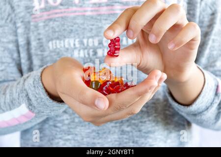 Una ragazza tiene in mano un orso gommoso, vuole mangiare dolci di gelatina. Primo piano. Concetto di delicatezza per bambini, cibo malsano, vitamine per bambini. Foto Stock