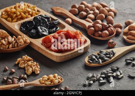 Varie varietà di noci, semi e frutta secca giacenti in cucchiai di legno e piatto su sfondo nero ardesia. Cibo sano. Alimentazione vegetariana Foto Stock