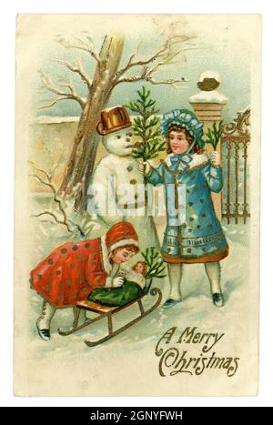 Biglietto d'auguri originale in rilievo per il Natale dell'epoca edoardiana britannica, le giovani ragazze con una bambola su una slitta nel loro giardino costruendo un pupazzo di neve, pubblicato da A.S.B. stampato in Germania, circa 1910 Foto Stock