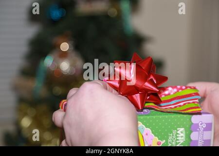 Apertura di un regalo dai colori vivaci che ha un arco rosso Su di esso davanti ad un albero di Natale Foto Stock