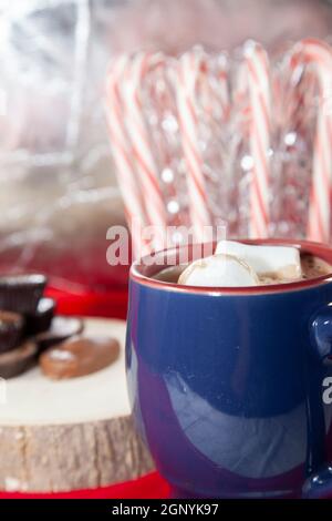 Tazza blu e rossa ripiena di cioccolata calda e due marshmallows, con cioccolatini su un ceppo di legno e canne candite sullo sfondo Foto Stock