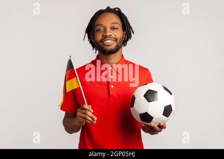 Uomo con un sorriso toothy indossando una T-shirt rossa casual, con bandiera tedesca e pallone da calcio bianco e nero classico e partita di avvistamento. Studio interno girato isolato su sfondo grigio. Foto Stock