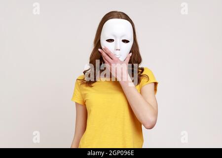 Donna anonima sconosciuta in giallo casual stile t-shirt coprendo il suo viso con maschera bianca, nascondendo personalità, cospirazione e privacy. Studio interno girato isolato su sfondo grigio. Foto Stock