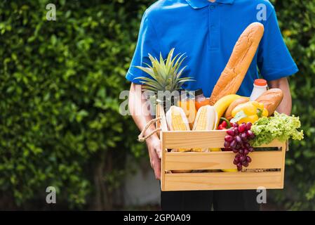 L'uomo agricolo asiatico indossa uniforme di consegna che tiene verdure fresche e frutta in scatola di legno di cassa in mani pronte dare al cliente raccolto organico Foto Stock