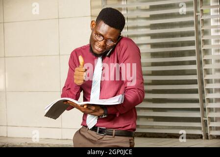 Il giovane africano impegnato fa due cose alla volta, parlando al telefono e gesturando mentre tiene il suo notebook. Un uomo interessato che tiene un telefono da t Foto Stock