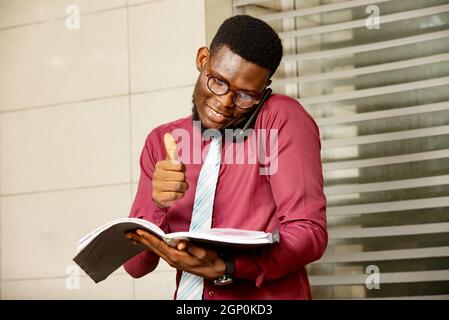 Il giovane africano impegnato fa due cose alla volta, parlando al telefono e gesturando mentre tiene il suo notebook. Un uomo interessato che tiene un telefono da t Foto Stock