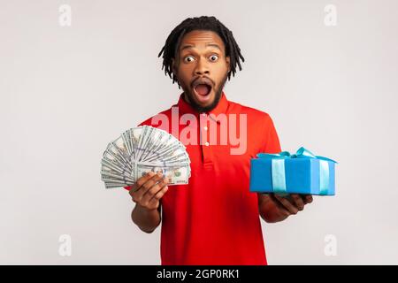 Stupito bell'uomo con dreadlocks indossare rosso casual stile T-shirt, tenendo molto denaro e presente scatola in mani, scioccato con bonus, omaggio. Studio interno girato isolato su sfondo grigio. Foto Stock