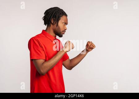 Ritratto del profilo arrabbiato di uomo con i doadlock che indossano la T-shirt di stile casual rosso, punzonatura, boxe con pugni aggrappati, essere pronto a combattere. Studio interno girato isolato su sfondo grigio. Foto Stock