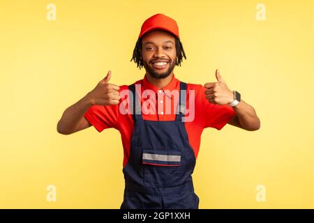 Ritratto del riparatore positivo indossando tute in piedi che mostrano i pollici in alto, guardando sorridendo la macchina fotografica, consiglia assistenza, manutenzione delle pulizie. Studio interno girato isolato su sfondo giallo. Foto Stock