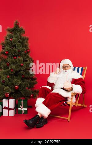 Babbo natale che tiene cocktail in cocco sulla sedia a sdraio vicino all'albero di natale su sfondo rosso Foto Stock