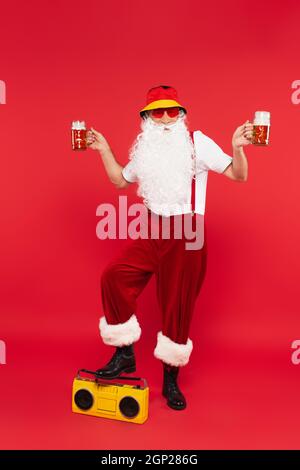 Babbo natale in occhiali da sole e cappello panama che tiene la birra vicino boombox su sfondo rosso Foto Stock