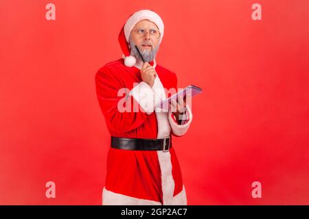 Ritratto di uomo anziano pensivo con barba grigia che indossa il costume di babbo natale tenendo in mano il taccuino, pensa ai regali per le vacanze di Natale. Studio interno girato isolato su sfondo rosso. Foto Stock