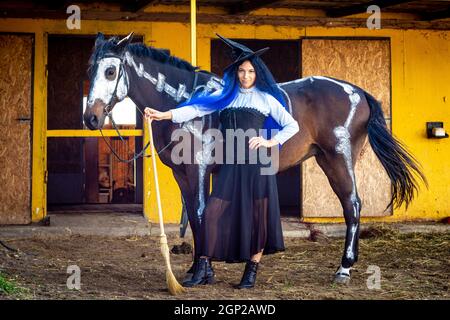 Una ragazza vestita come una strega si alza con una scopa vicino ad un cavallo su cui è disegnato uno scheletro Foto Stock