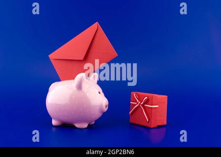 Busta rossa festiva inserita in una banca rosa piggy accanto a una casella presente su sfondo blu con copia spazio Foto Stock