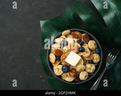 Cibo alla moda - pancake cereali serviti burro e mirtilli. Mucchio di mini frittelle di cereali in giro su foglia tropicale monsterra su sfondo scuro. COP Foto Stock