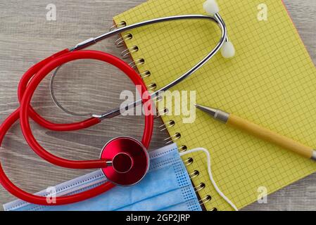 Tema medico ancora vita con uno stetoscopio rosso colorato e. maschera chirurgica posizionata sul lato del filo legato blocco note e penna visti come una posa piatta Foto Stock