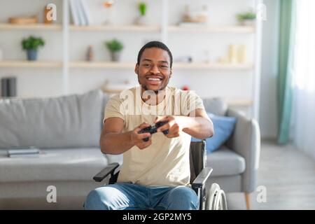 Gioiosa paraplegica nera in sedia a rotelle che si diverte a divertirti con il gioco su playstation a casa. Giocatore maschio disabilitato con controller che gioca al computer. Lockdo Foto Stock