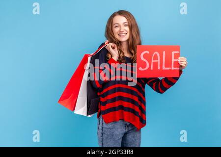 Donna allegra vestendo a righe stile casual maglione, vantando con acquisti che tengono carta shopping borse e carta di vendita, grandi sconti per le vacanze. Studio interno girato isolato su sfondo blu. Foto Stock