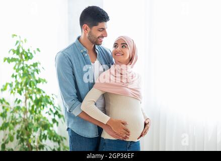 Amando gli sposi arabi coccolarsi e godersi la futura parenting, l'uomo abbracciare la moglie e toccare il pancino, sorridendo l'un l'altro, in piedi all'interno di casa, f Foto Stock