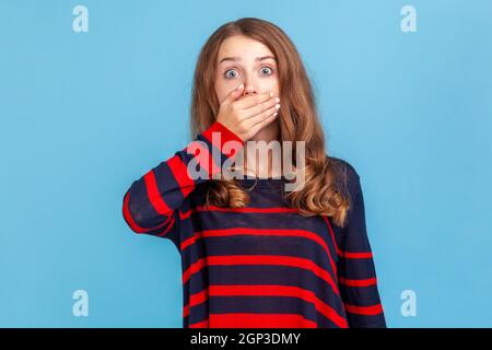 Non dire! Ritratto di donna spaventata in maglia casual a righe che copre la bocca con la mano spaventato a parlare, mantenendo segreto, guardando sconvolto preoccupato. Studio interno girato isolato su sfondo blu. Foto Stock