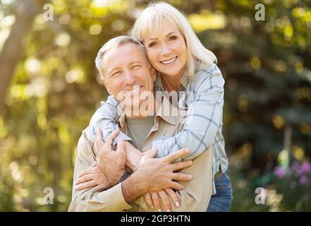 Uomo e donna anziani felici che trascorrono del tempo insieme, abbracciando, riposando in giardino al sole della giornata autunnale all'aperto e sorridendo alla macchina fotografica, spazio copia Foto Stock