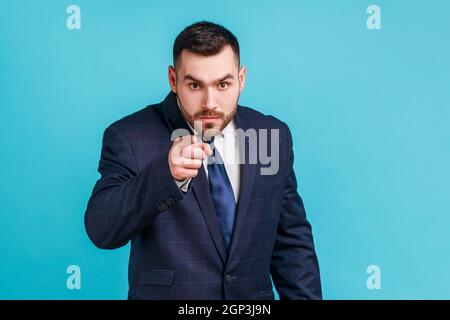 Uomo serio con binched che indossa un abito ufficiale con un dito puntato, che indica la direzione della fotocamera e che ha un aspetto infastidito, facendo la scelta. Studio interno girato isolato su sfondo blu. Foto Stock