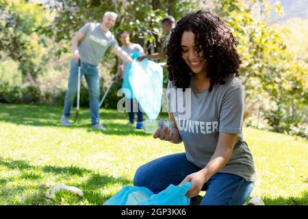 Sorridente donna afroamericana che tiene sacco di rifiuti, raccogliendo rifiuti di plastica in campo con gli amici Foto Stock