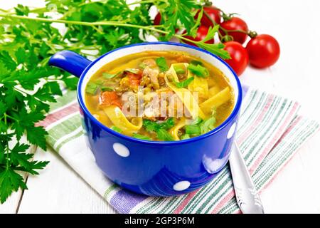 Zuppa con carne, pomodori, verdure, lenticchie di fagioli e tagliatelle in una ciotola blu su asciugamano, prezzemolo e un cucchiaio su sfondo di legno Foto Stock
