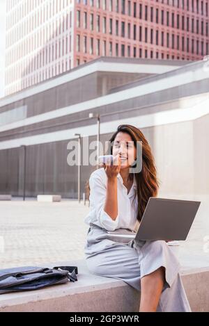 ritratto verticale di una giovane ed elegante donna indiana. Sta sedendo vicino all'edificio dell'ufficio e sta lasciando un messaggio vocale con il telefono Foto Stock