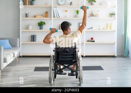 Vista posteriore del tipo nero praplegico in sedia a rotelle che si allenano con i manubri a casa, a tutta lunghezza. Disabili Afro uomo esercizio, facendo fisioterapia indo Foto Stock