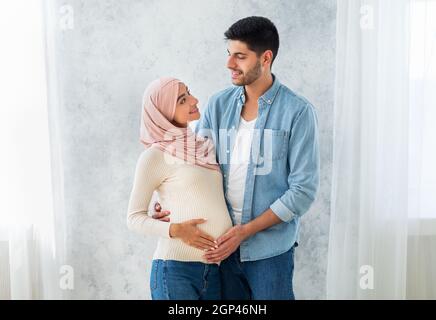 Felici genitori-per-essere. Felice coppia musulmana in attesa del bambino, uomo che abbraccia la moglie e tocca il pancino, in piedi all'interno di casa, spazio libero. Spus islamico Foto Stock