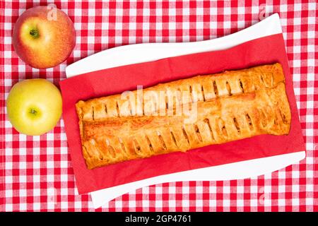 Apple roll servito. Vista dall'alto di uno strudel di mele fresco cotto in casa con due mele fresche su un panno rosso da picnic o un tovagliolo. Concetto di torta e pasticceria. Spin Foto Stock