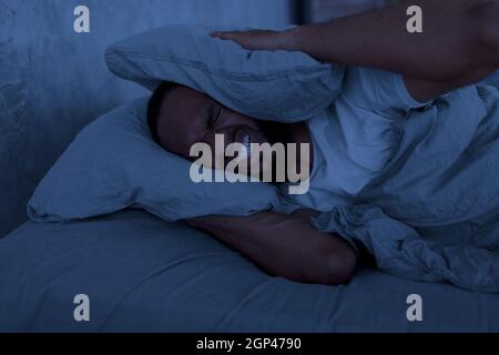 Stress, Insonnia, collera Cocnept. Ritratto del giovane afroamericano arrabbiato sdraiato a letto di notte e coprendo le sue orecchie di cuscino, sofferente di Foto Stock
