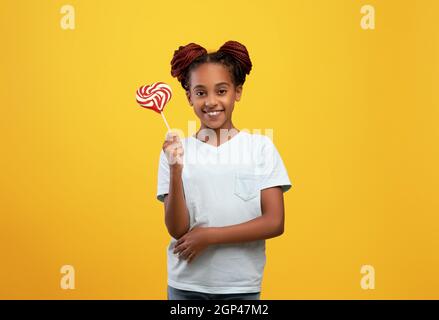 Carino afroamericana ragazza adolescente tenendo colorato cuore a forma di caramella e sorridendo alla macchina fotografica, allegra ragazza nera teen in t-shirt bianca che posa sul giallo Foto Stock
