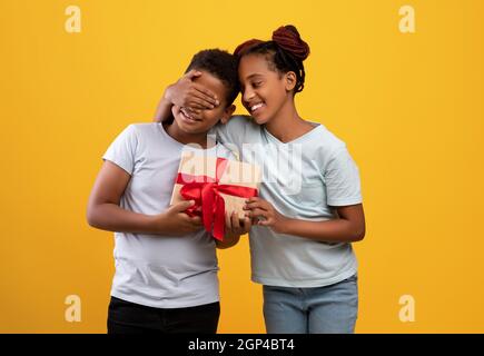 Felice teen ragazza nera amorevole sorella saluto suo fratello con compleanno, chiudendo gli occhi del ragazzo con la palma e dando scatola regalo, sfondo giallo studio. Afro Foto Stock