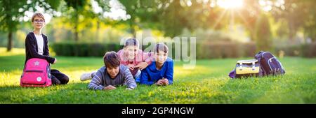 Bambini con zaini in piedi nel parco vicino a scuola. Alunni con libri e zaini all'aperto giacenti sull'erba Foto Stock