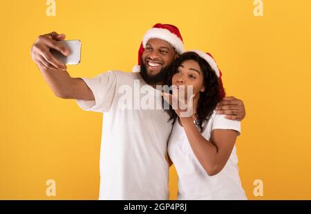 Allegro sposi afroamericani in Babbo Natale con selfie su smartphone, donna che soffia aria bacio cellulare fotocamera, in posa su studio giallo backgr Foto Stock