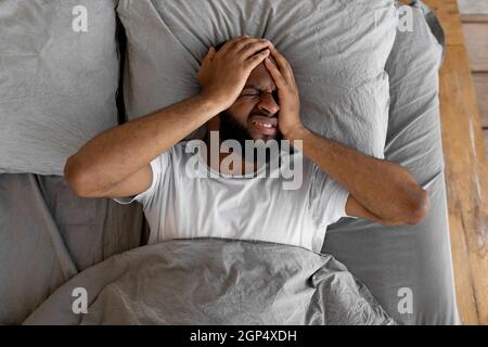 Vista dall'alto del giovane uomo nero che soffre di mal di testa o emicrania dopo il risveglio di mattina. Ragazzo stressato sdraiato a letto con viso doloroso Foto Stock