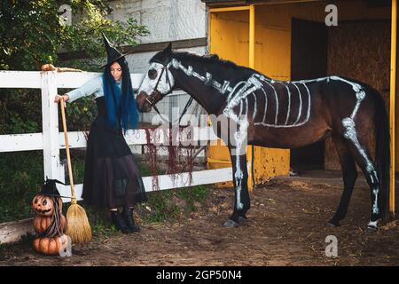 Una ragazza vestita da strega si trova accanto alla recinzione del Corral, e accanto ad essa si trova un cavallo con uno scheletro dipinto Foto Stock