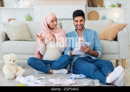 I coniugi arabi incinta eccitati si preparano per il parto, facendo lista di controllo delle necessità, seduti insieme sul pavimento in salotto con il vestito del bambino Foto Stock