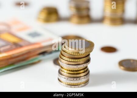 Monete in euro in colonna su tavola bianca con un mucchio di banconote in background. Contanti impilati in pile da vicino. Concetto di ricchezza ed economico Foto Stock