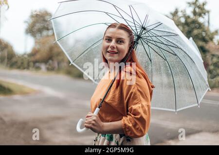 Bella donna contented in abiti luminosi sotto un ombrello trasparente in caso di maltempo. Foto Stock