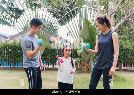 Giovane madre, padre e figlia asiatica fare esercizio insieme a dumbbells è divertimento all'aperto nella natura un campo giardino parco. Buon bambino di famiglia Foto Stock