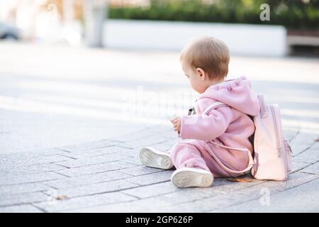 Divertente bambina 1 anno di età indossare elegante tuta rosa sport e zaino seduta su strada su sfondo urbano. Bambino piccolo carino con c trendy Foto Stock